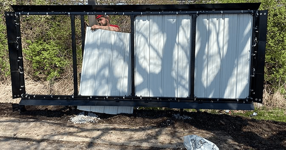 A man wearing a red shirt and a hard hat changes out panels on a large metal billboard structure near several trees.
