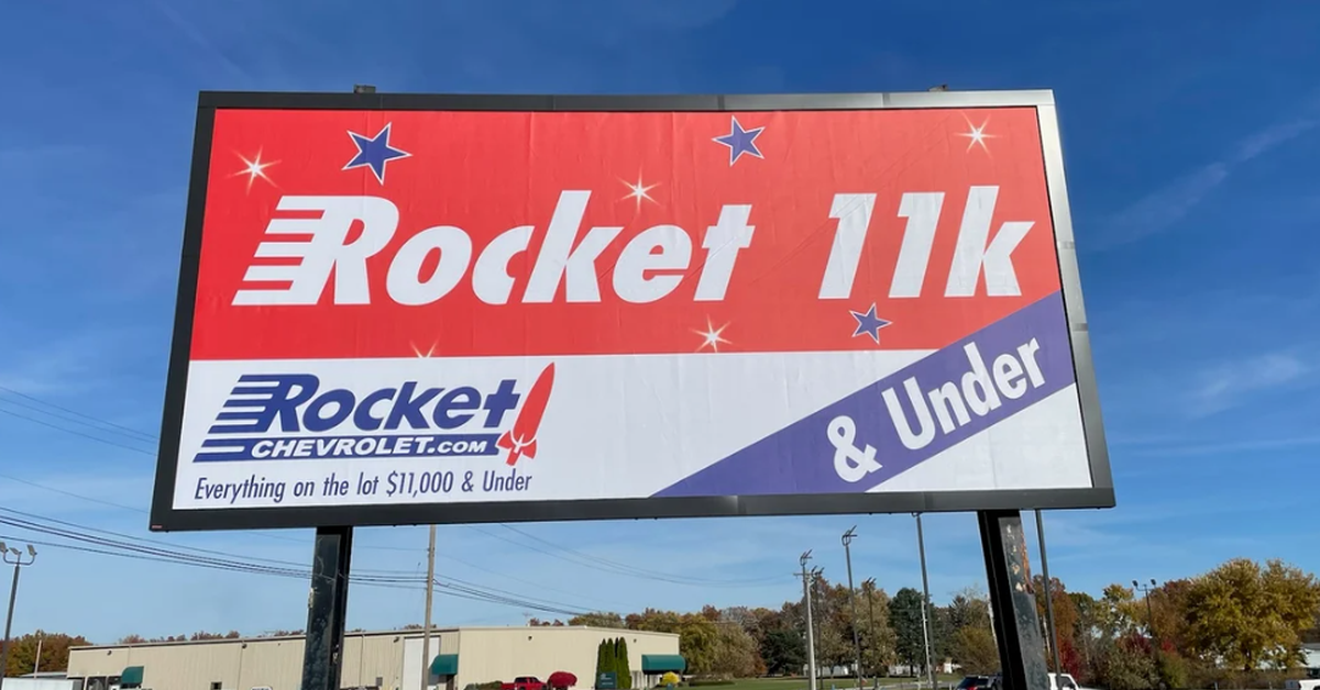 A freestanding billboard banner with the words "Rocket 11K" in an open field near a warehouse and parking lot.