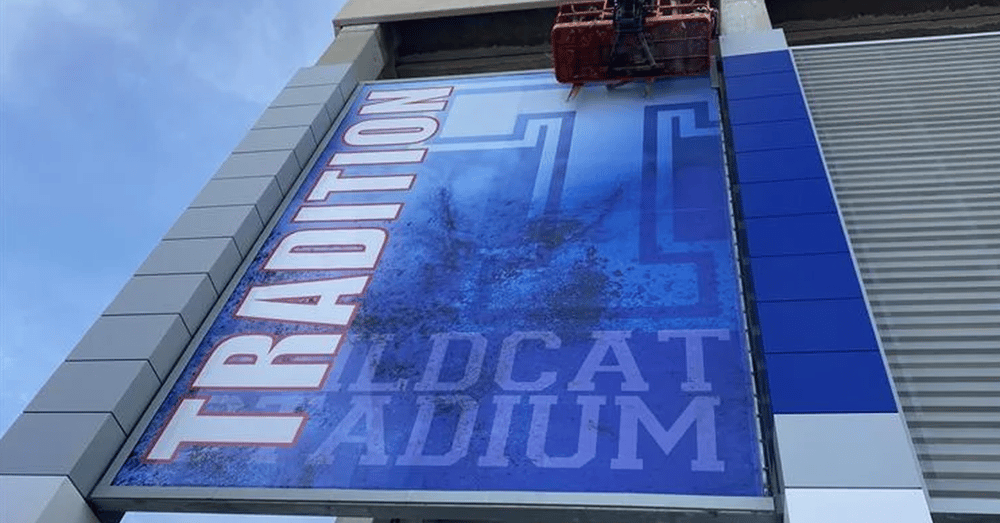 A low-angle view of a blue banner hanging on a stadium while an elevated platform works near the upper edge.