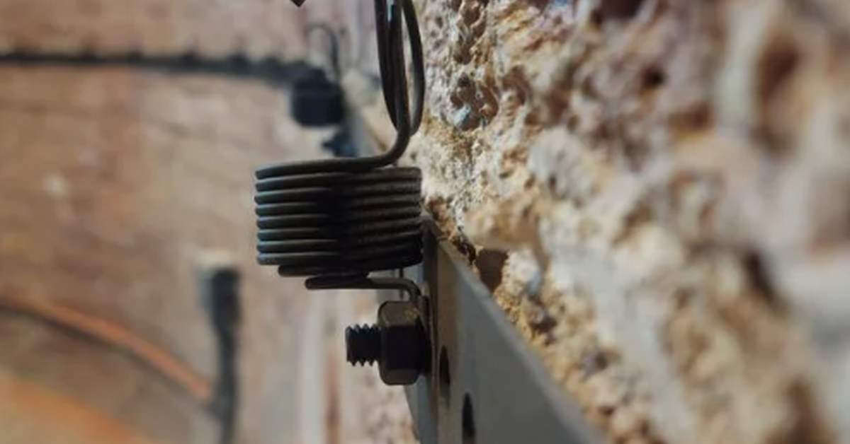 A close-up of a metal spring and hook fastened to a textured brick wall beside a perforated metal strip.