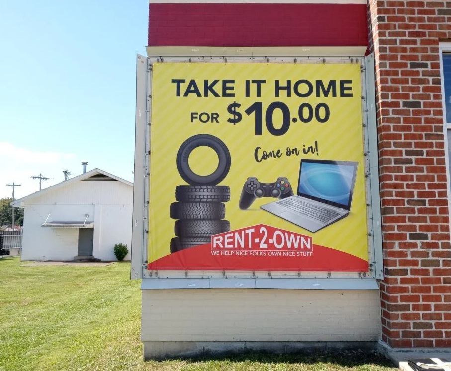 A yellow Rent-2-Own sign on a brick building advertising tires, a game controller, and a laptop for ten dollars.