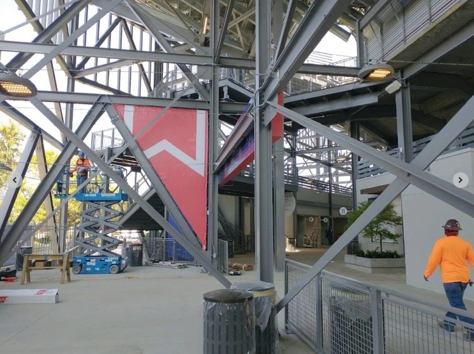 Workers and equipment beneath metal stadium beams, with a lift, ladder, signage, and walkways visible.