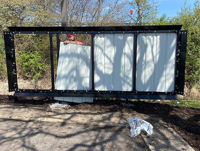 A man wearing a red shirt and a hard hat changes out panels on a large metal billboard structure near several trees.