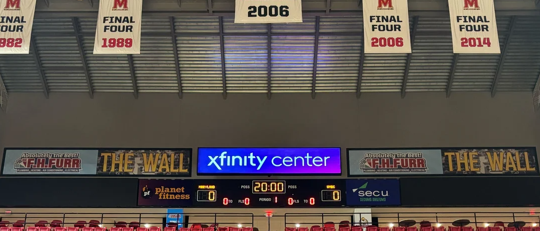 An indoor arena scoreboard below banners, sponsor signs, red seats, and a bright Xfinity Center sign.