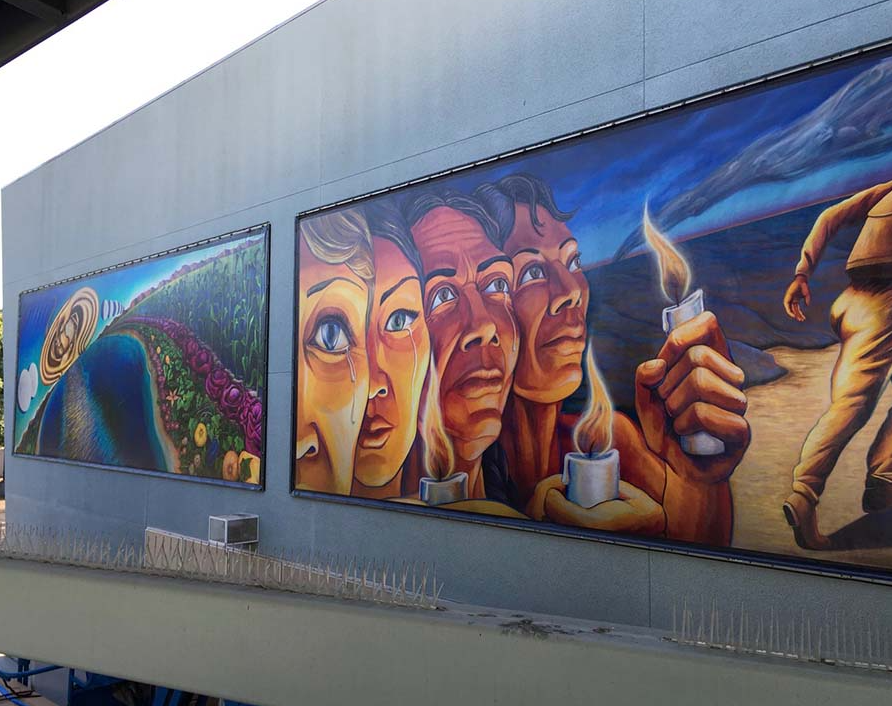 Two large murals displayed on a gray building, including painted faces, hands holding candles, and a landscape.