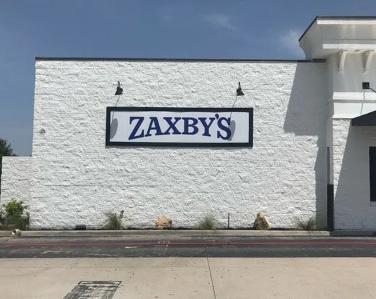 A Zaxby's wall sign mounted on a white textured building exterior above a sidewalk and curb on a sunny day.