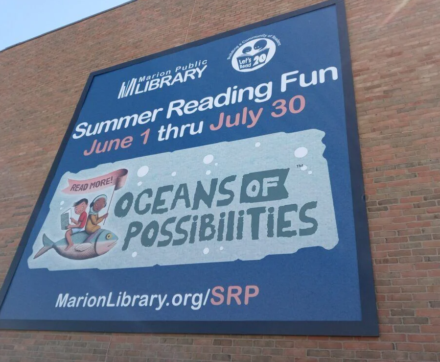 A large blue library banner mounted on a brick building promoting a summer reading program with illustrated children.