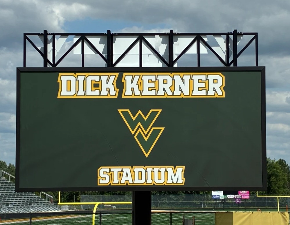 A large green Dick Kerner Stadium sign with a yellow WV logo mounted above a sports field under a cloudy sky.