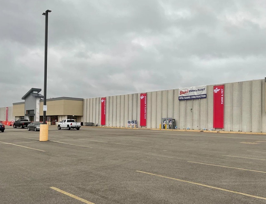 A large commercial building with red vertical banners, parked vehicles, and an empty parking lot under cloudy skies.