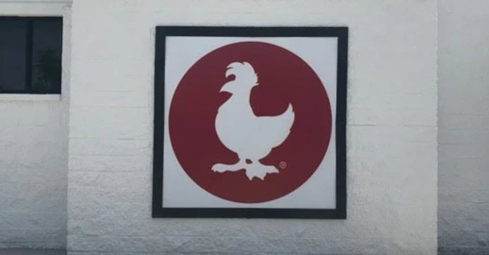A framed sign featuring a white chicken silhouette inside a red circle mounted on a white brick exterior wall.
