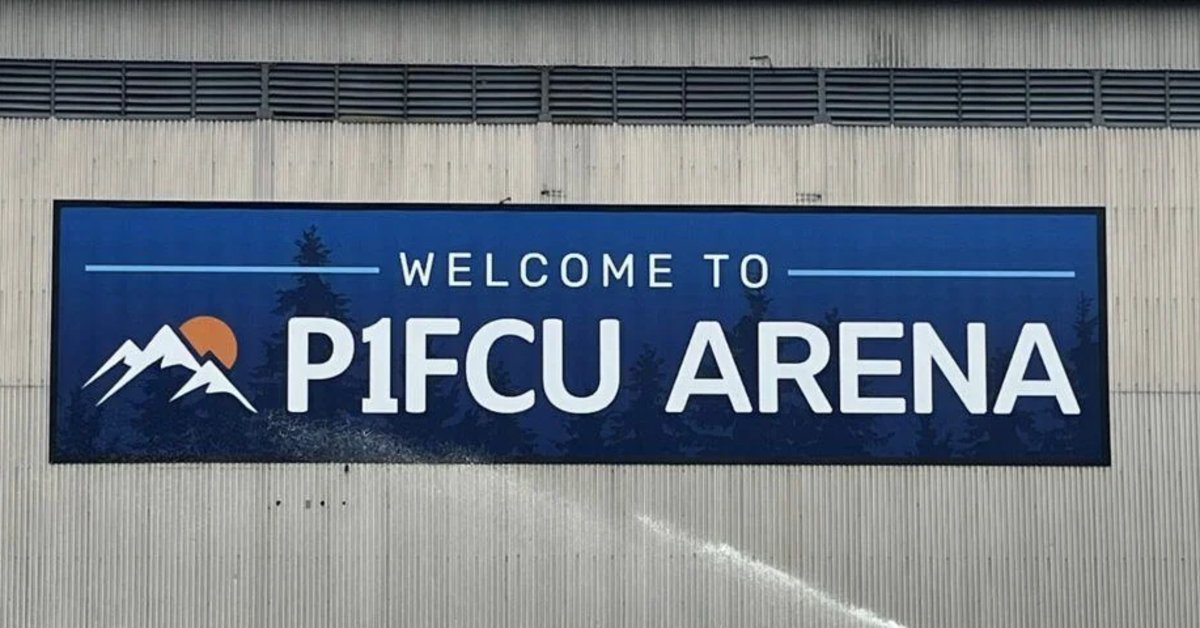A large blue exterior sign reading “Welcome to P1FCU Arena,” featuring a mountain logo and forest silhouette graphics, mounted on an exterior wall.
