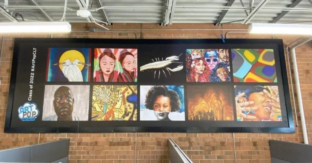 A large indoor art installation displaying a grid of colorful student artwork, including portraits, abstract designs, and cultural themes, mounted on a brick wall above a stairway.