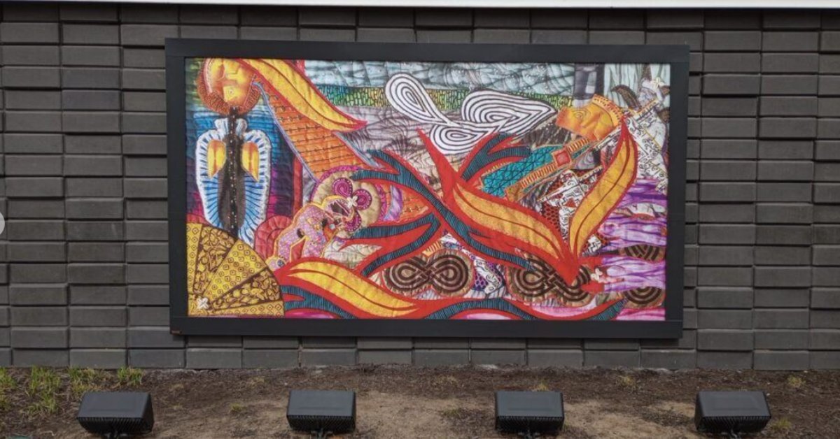 Framed outdoor art installation showcasing a colorful abstract mural mounted on a modern building facade with landscape lighting.