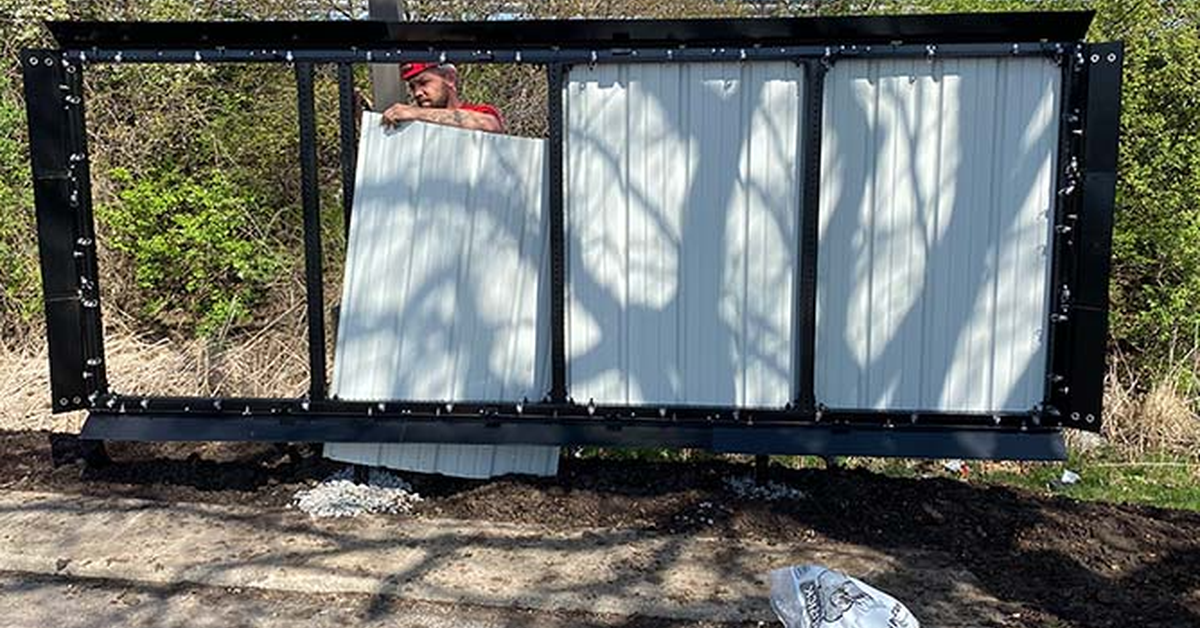 A man wearing a red shirt and a hard hat changes out panels on a large metal billboard structure near several trees.