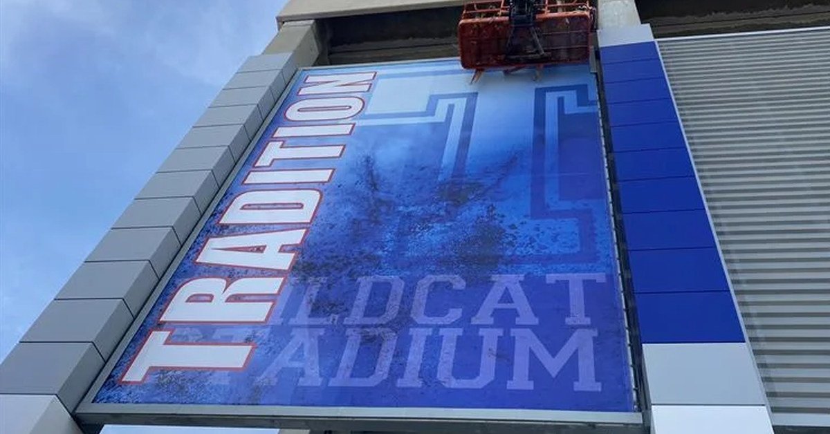 A low-angle view of a blue banner hanging on a stadium while an elevated platform works near the upper edge.