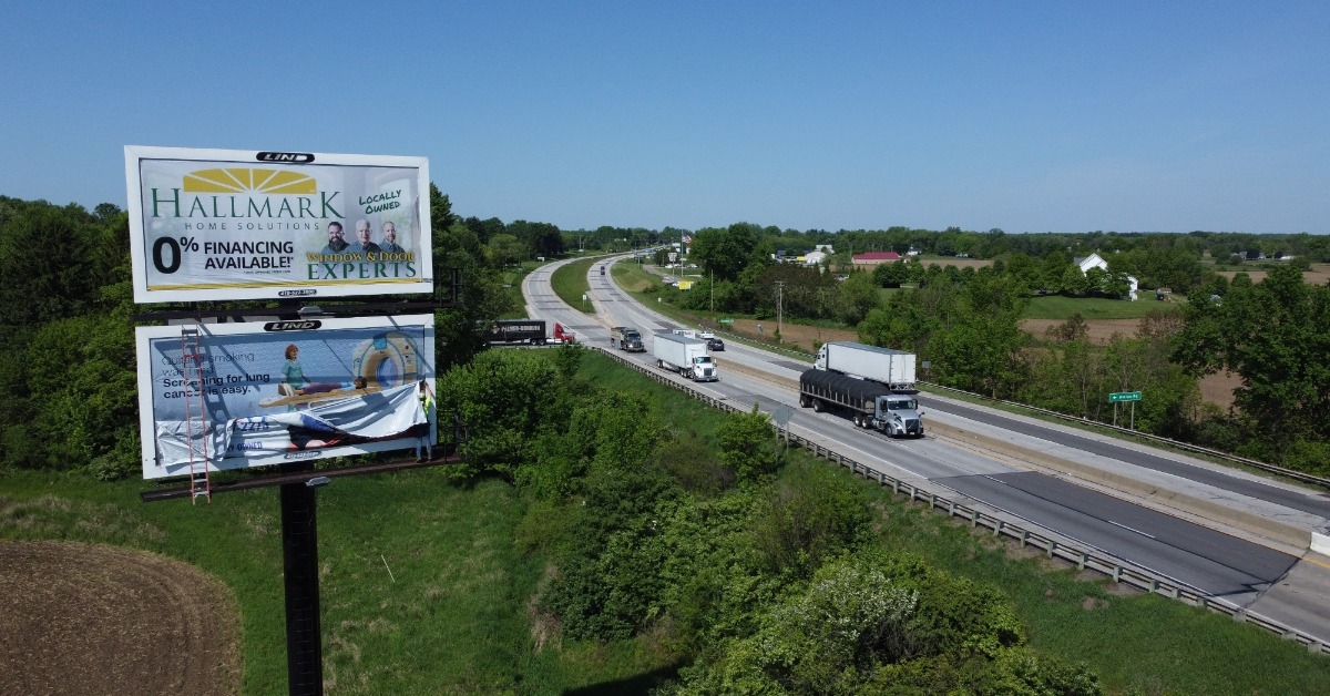 Multiple billboards sitting in a field next to a highway, against a bight blue sky