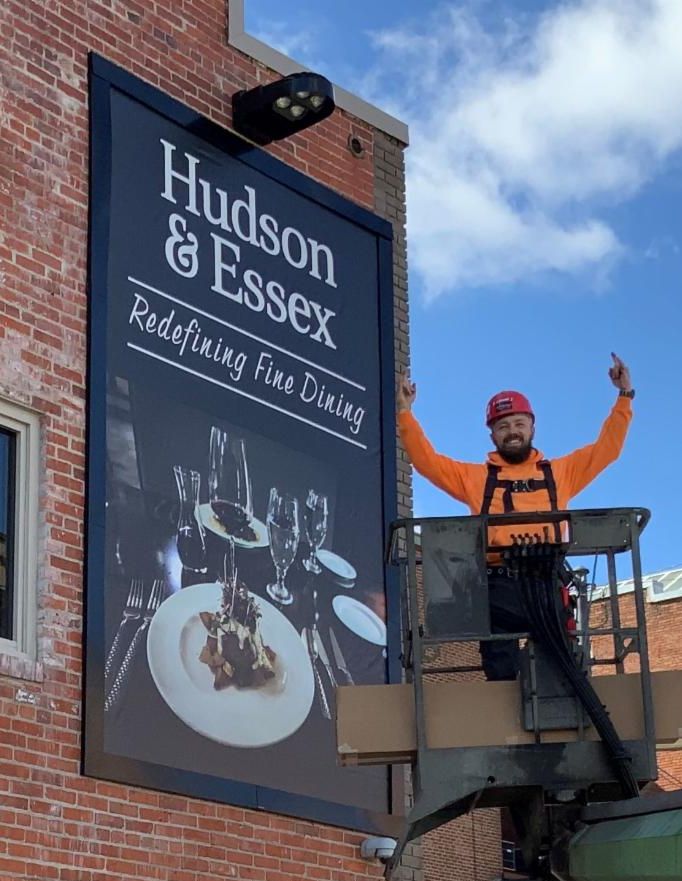 A Lind SignSpring Product – BannerFrameCLASSIC With Covers for Hudson & Essex on an exterior brick wall with the sign installation man with his hands raised jubilantly