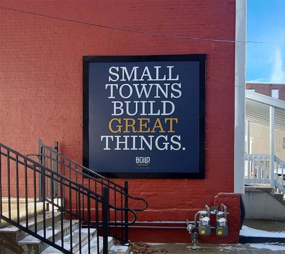 A Lind SignSpring Product – BannerFrameCLASSIC With Covers for a square banner with the text “Small Towns Build Great Things” on an exterior brick wall